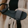 Sort KIRA-sandal med ankelrem og tyk sål, holdt i hånden foran en beige frakke.